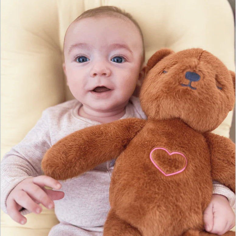 Baby holding a brown teddy bear with a heart on a beige cushion, featuring Lululeo branding.