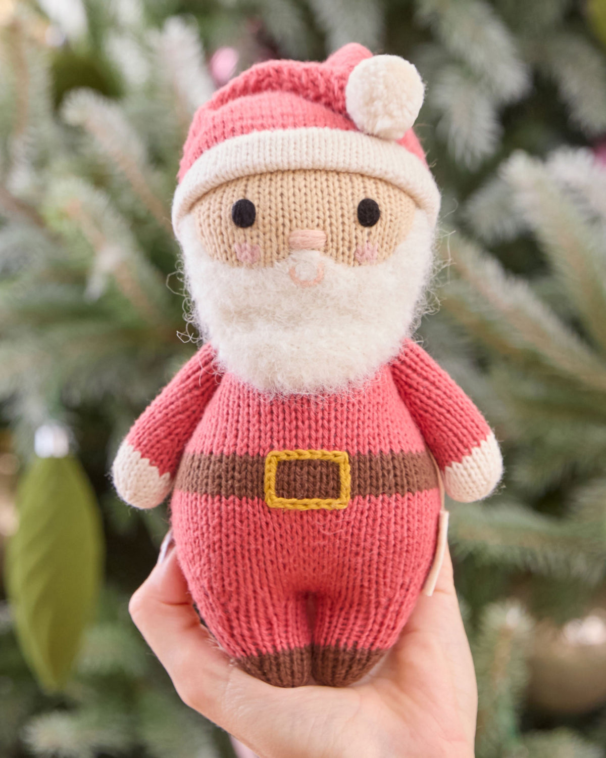 Soft Baby Santa plush toy by Cuddle + Kind with red outfit, holding a tiny gift, perfect for Christmas cuddles and holiday decor.