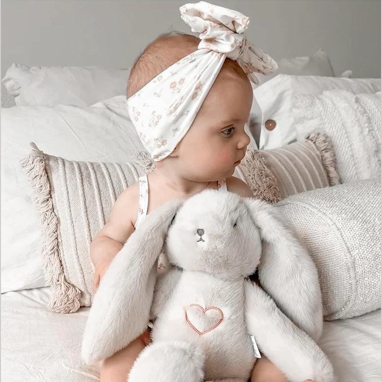 Baby holding a plush bunny toy on a bed with light-colored pillows.